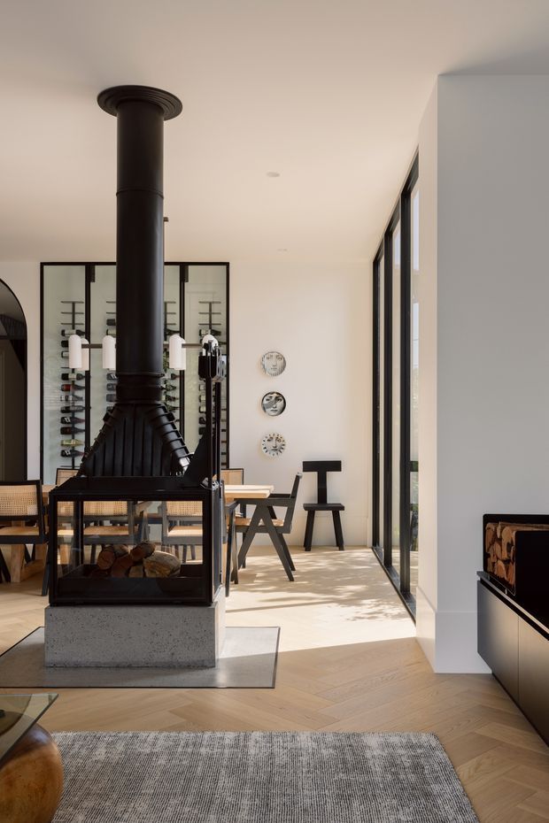 A bright, refined open-plan living and dining area with a prominent black freestanding wood-burning fireplace and flue on a light stone/concrete plinth. The room features pale herringbone timber flooring, white walls, full-height black-framed glazing, a timber dining table with black chairs, built-in dark cabinetry, and a large neutral rug. Decorative shelving and wall-mounted plates add visual interest, while sunlight creates patterned shadows across the floor.