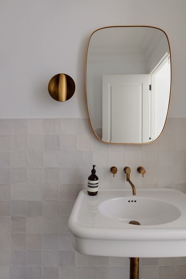 A refined bathroom vanity vignette featuring a white pedestal-style basin, pale square wall tiles, a brass wall-mounted tap set, an organically shaped brass-framed mirror, and a sculptural brass wall light. The mirror reflects a white paneled door and detailed ceiling trim. The palette is soft white and grey with warm brass accents.