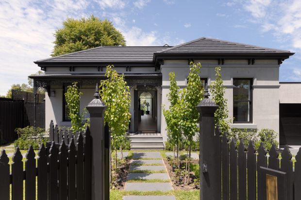 Front exterior view of a single-storey rendered home with a low hipped tiled roof, covered entry porch, symmetrical facade, tall windows, and a landscaped path leading through a dark picket-style front fence and gate.