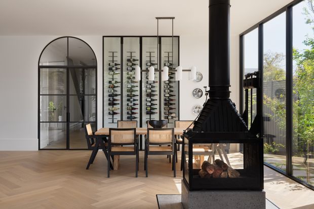 Bright contemporary dining area with herringbone timber flooring, a large black freestanding fireplace, floor-to-ceiling glazing, and a black-framed arched internal glass door. A timber dining table and chairs sit in front of a decorative wine-bottle display, with greenery visible outside.
