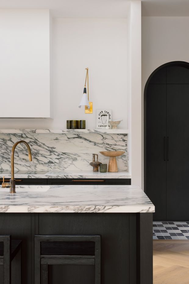 A refined kitchen interior featuring a dark timber island with seating, white cabinetry, dramatic white-and-grey veined marble benchtops and backsplash, brass tapware, and a wall-mounted pendant light. A black arched doorway leads to a tiled adjoining space, while warm timber flooring adds contrast.