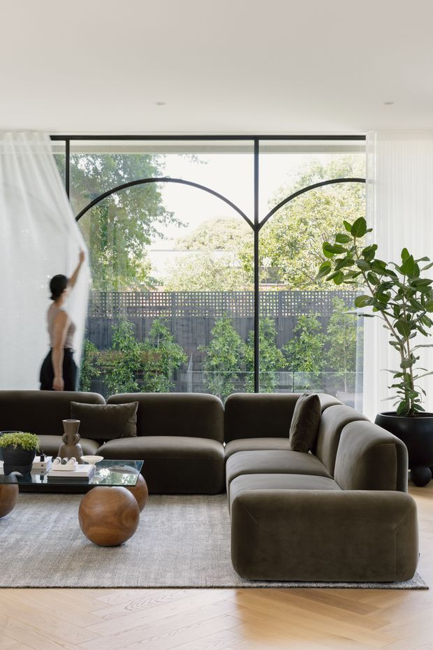 Bright contemporary living room with a large black-framed glazed wall overlooking a planted courtyard or garden. A deep olive-brown sectional sofa sits on a large neutral rug, accompanied by a rounded wooden coffee table, sculptural ceramic decor, and a tall indoor plant. White walls and sheer curtains create an airy setting, while the patterned timber parquet floor adds warmth.