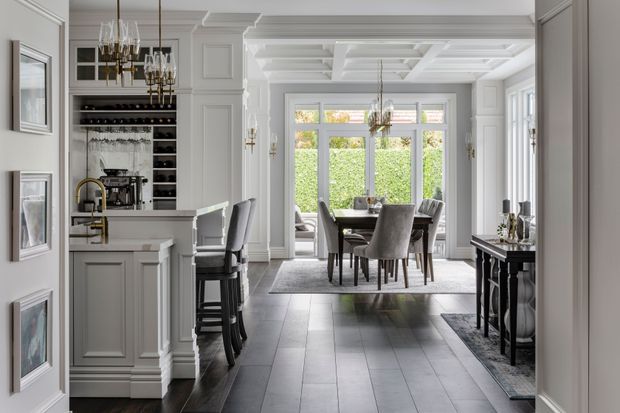 Elegant open-plan interior combining a compact kitchen and bar area with a formal dining room. The space features white painted panelled cabinetry and walls, dark timber flooring, a coffered ceiling, framed artwork, a dark sideboard, upholstered dining chairs, and large glazed doors and windows opening toward a leafy garden.