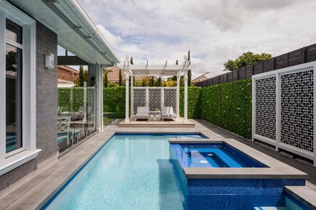 A contemporary residential backyard with a long swimming pool and attached spa, bordered by dark tiled paving and clipped green hedges. The space includes a white pergola with outdoor seating, decorative patterned privacy screens, and a glazed house facade along one side.