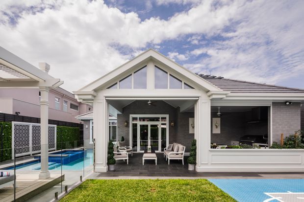 Rear exterior view of a refined single-storey gable-roof home with a covered outdoor entertaining area, glazed doors, swimming pool, and an open outdoor kitchen or servery. The facade uses pale painted finishes with grey masonry, white trim, and a tiled roof.