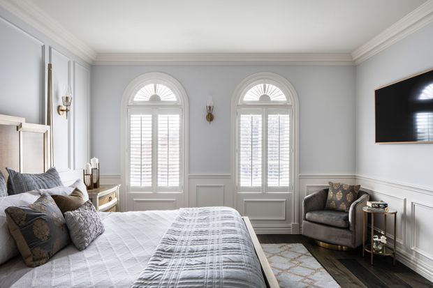Elegant bedroom with two tall arched windows fitted with white plantation shutters, pale grey wall panelling and wainscoting, ornate crown moulding, dark timber flooring, and a large upholstered bed. A grey armchair, side tables, wall sconces, and wall-mounted television complete the refined interior.