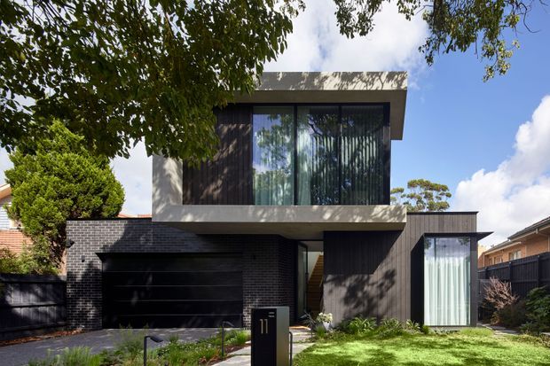 Contemporary multi-level urban residence with a dark brick and vertical timber-clad facade, projecting upper floor, large glazed windows, and a flat roof. The home includes an integrated single garage, landscaped front garden, and a recessed entry walkway.