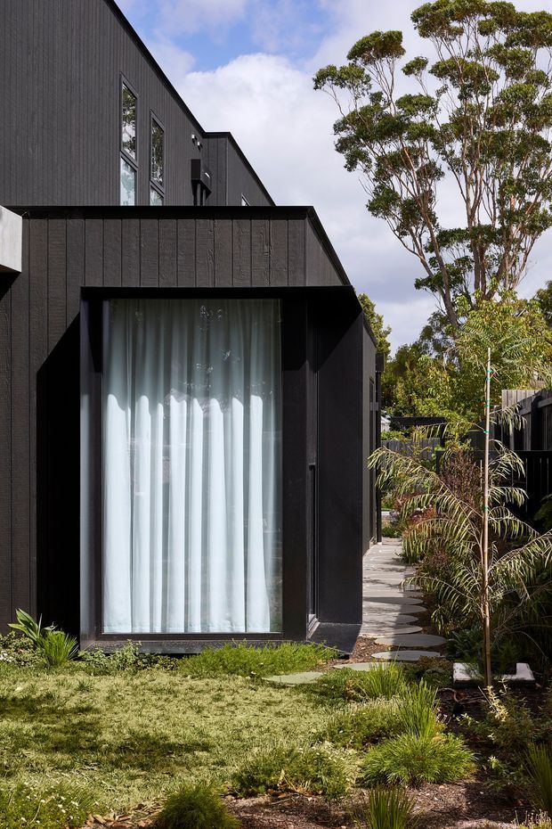 A contemporary multi-level residential exterior clad in dark vertical textured panels, featuring a large floor-to-ceiling glazed opening with sheer curtains. A narrow stepping-stone path runs through a landscaped garden beside the house, with mature trees and layered planting surrounding the building.