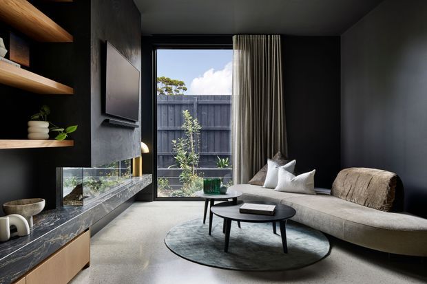 A moody contemporary living room with dark charcoal walls and built-in shelving, a long stone-clad media and fireplace wall, floor-to-ceiling glazing with curtains, and a curved neutral sofa arranged around nested black coffee tables on a round rug. A small planted courtyard is visible beyond the glazing.