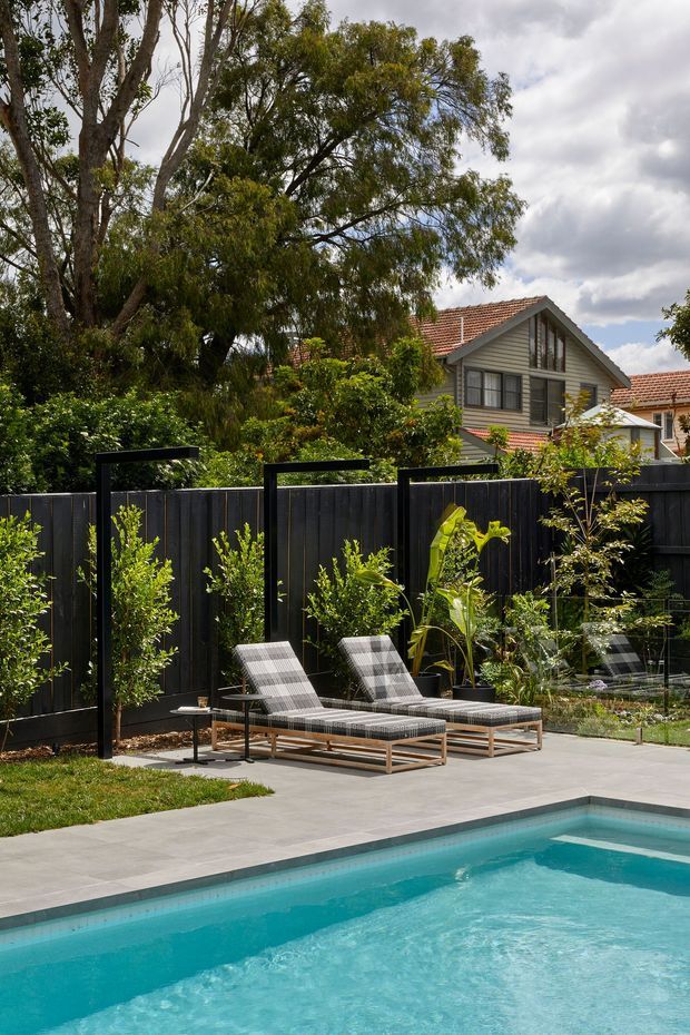 A landscaped residential backyard with a turquoise swimming pool in the foreground, large pale concrete paving, timber-framed reclining lounge chairs, and a black vertical paling fence. The garden includes established trees, shrubs, and tropical planting, with a traditional gabled weatherboard house visible beyond the fence.