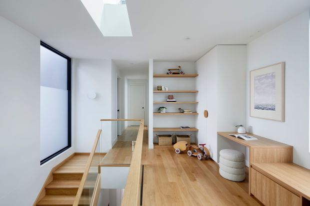 Bright, minimalist upper-level hallway and landing with warm timber flooring, a timber stair balustrade, built-in open shelving, and a low timber cabinet. A skylight and large window provide abundant natural light, while children's toys and a small upholstered stool add a lived-in family character.