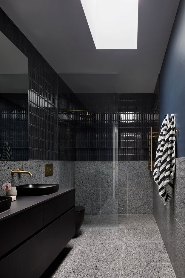 A dramatic contemporary bathroom with dark charcoal and black finishes. The room features a floating black vanity with a dark vessel basin, brass tapware and hardware, grey speckled stone-look wall and floor tiles, glossy black vertical wall tiles, a large mirror, a skylight, and a black-and-white striped towel hanging on a brass rail.