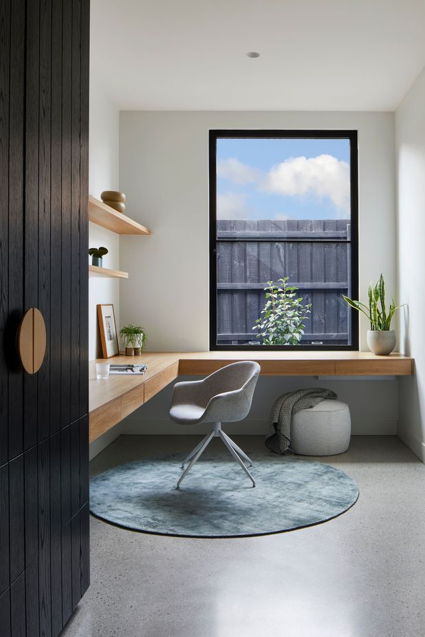 A compact, thoughtfully designed home office nook with a built-in timber desk wrapping around the corner beneath a large black-framed window. Dark vertical timber wall panelling and matching cabinetry contrast with pale walls and a polished concrete floor. Open timber shelves display small decorative objects and plants, while a grey swivel chair, round blue-grey rug, and upholstered ottoman create a calm, contemporary workspace.