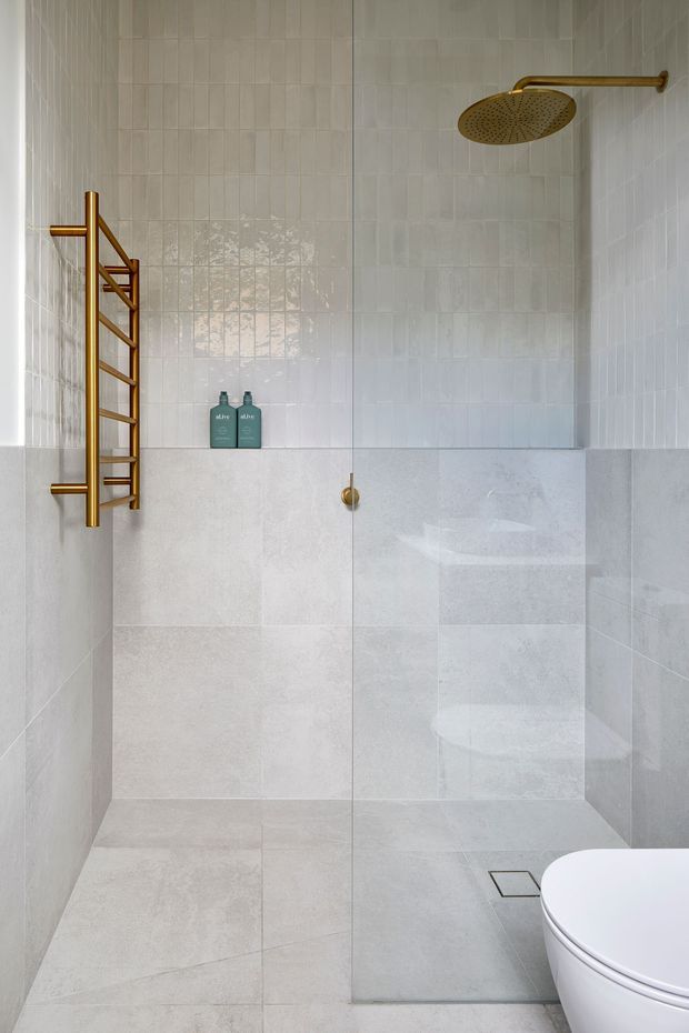Contemporary bathroom shower with full-height pale grey and white tiled walls, large-format floor tiles, a frameless glass shower screen, and brushed brass fixtures including a rain shower and heated towel rail. A partial white toilet is visible in the foreground. The space is bright, clean, and minimal with subtle tile texture.