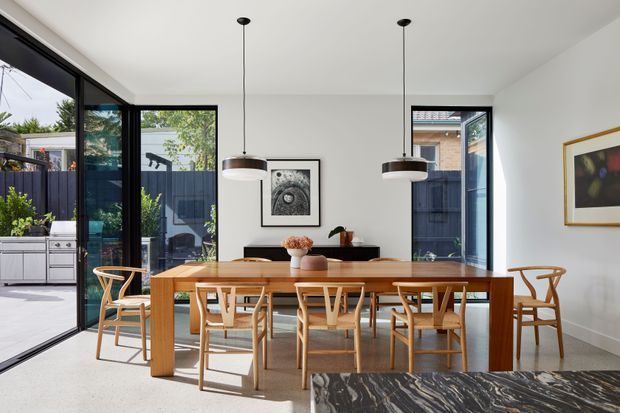 Bright contemporary dining room with a long solid-wood dining table and matching timber chairs, two suspended pendant lights, white walls, large black-framed glazed doors, and views to a private outdoor courtyard. A dark marble or stone surface is visible in the foreground, with framed artwork and a compact sideboard adding restrained decoration.