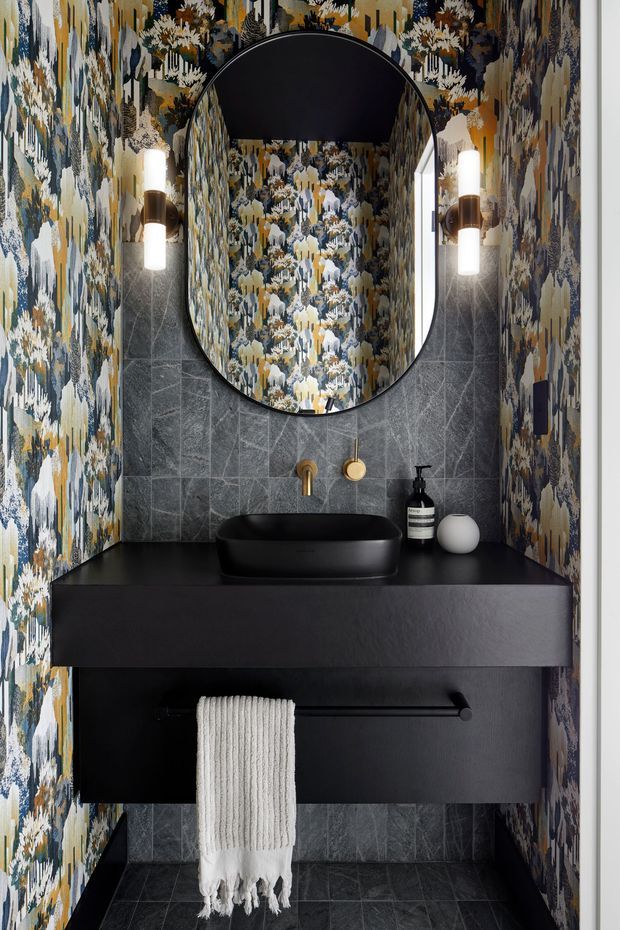 A compact, dramatically styled bathroom vanity area with a black vessel basin on a floating black vanity, dark grey stone-look tiled wall and floor, and bold botanical-patterned wallpaper on the side walls and inside the arched mirror reflection. An oversized rounded-arch mirror, cylindrical wall sconces, brass tapware, and a white hand towel complete the refined high-contrast scheme.