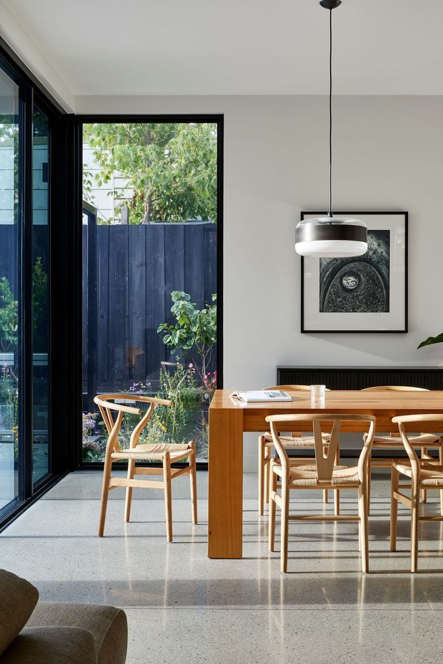 A bright, refined dining area with a warm timber dining table and woven-seat timber chairs, positioned beside full-height black-framed glazing opening to a planted courtyard. The room has a smooth white ceiling and walls, a polished light terrazzo floor, a dark built-in sideboard, a sculptural pendant light, and framed monochrome artwork. A partial sofa is visible in the foreground.