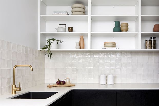 Bright contemporary kitchen with white open shelving, glossy white tiled splashback, white benchtops, dark lower cabinetry, and a brass gooseneck tap over a black sink. The shelves display neutral-toned ceramics and small decorative objects, with a trailing plant adding an organic touch.