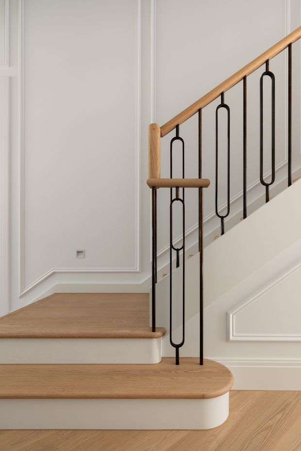 Close-up architectural photograph of a refined staircase with rounded light timber treads and handrail, white-painted risers and walls, decorative wall panelling, and slender black metal balusters featuring elongated loop motifs. The composition highlights the stair detailing and warm neutral material palette.