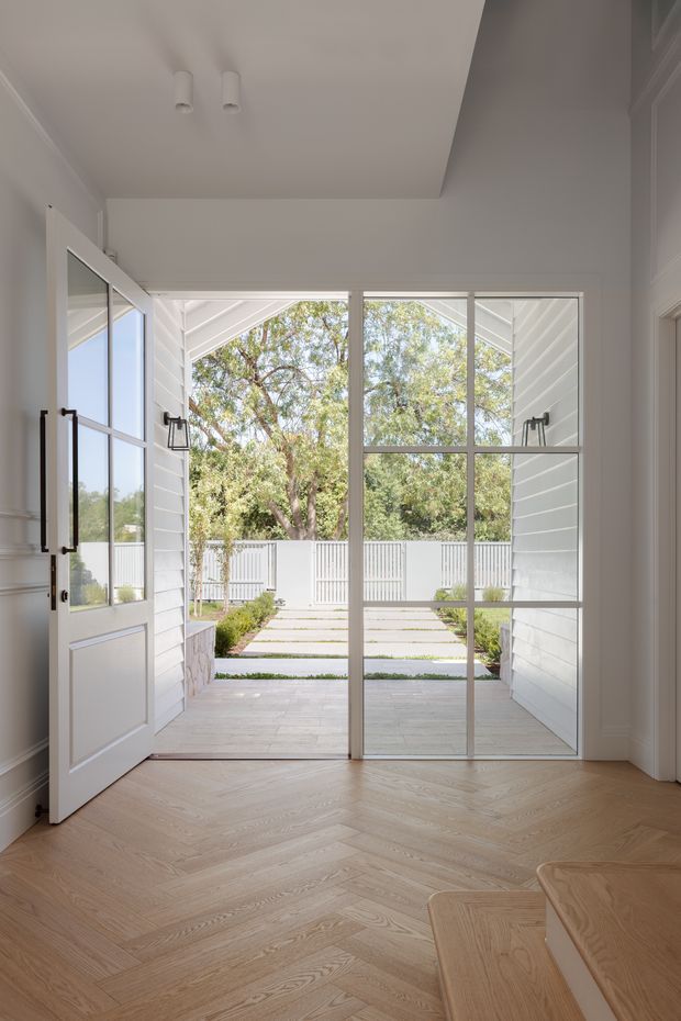 Bright residential entryway viewed from inside, featuring a white paneled hinged door, large steel-framed glazed doors, white walls and ceiling, and a light timber herringbone floor. The doors open to a paved courtyard or terrace with white horizontal-clad walls, garden planting, and a white balustrade. A small portion of built-in or adjacent timber cabinetry is visible at the lower right.