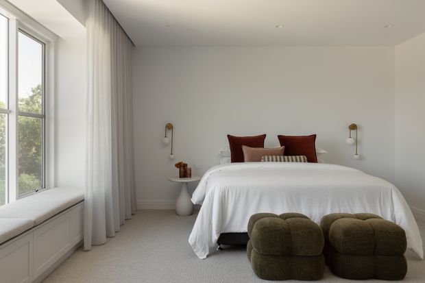 A serene, minimalist bedroom with a low-profile bed dressed in white bedding, dark rust-brown and neutral pillows, and two rounded olive-brown upholstered ottomans at the foot. Full-height sheer curtains frame a large window with a built-in window seat and cabinetry below. White walls, recessed ceiling lights, carpeted flooring, and small brass wall-mounted reading lights create a calm, refined interior.