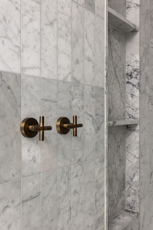 Close-up of a refined bathroom shower wall clad in pale grey-white marble tiles with prominent natural veining. Two wall-mounted brushed bronze controls provide contrast, while a recessed marble-lined shower niche with shelves is visible on the right.