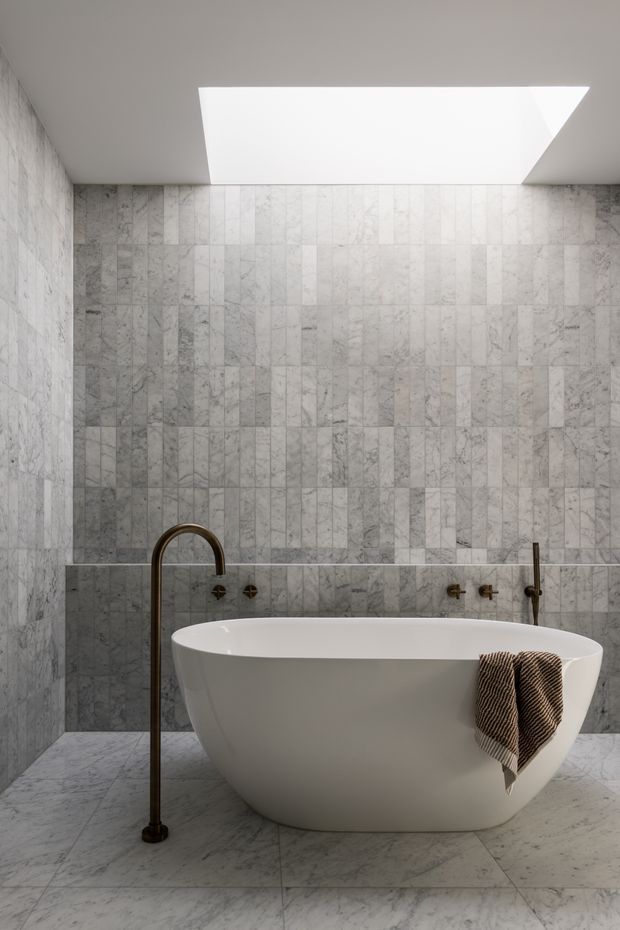 A refined minimalist bathroom clad throughout in pale grey-white veined marble or stone tiles. A freestanding white oval bathtub sits centrally beneath a large rectangular skylight, with dark bronze freestanding and wall-mounted bath fittings. The room has a calm, monochromatic palette and strong natural light.
