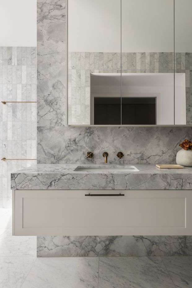 A refined contemporary bathroom vanity featuring extensive grey-veined marble or stone surfaces, pale grey wall and floor tiles, a floating light-grey vanity with a dark metal handle, integrated basin, brass-toned wall-mounted tapware, and mirrored wall cabinetry. A narrow marble ledge and restrained decorative styling create a calm, luxurious composition.