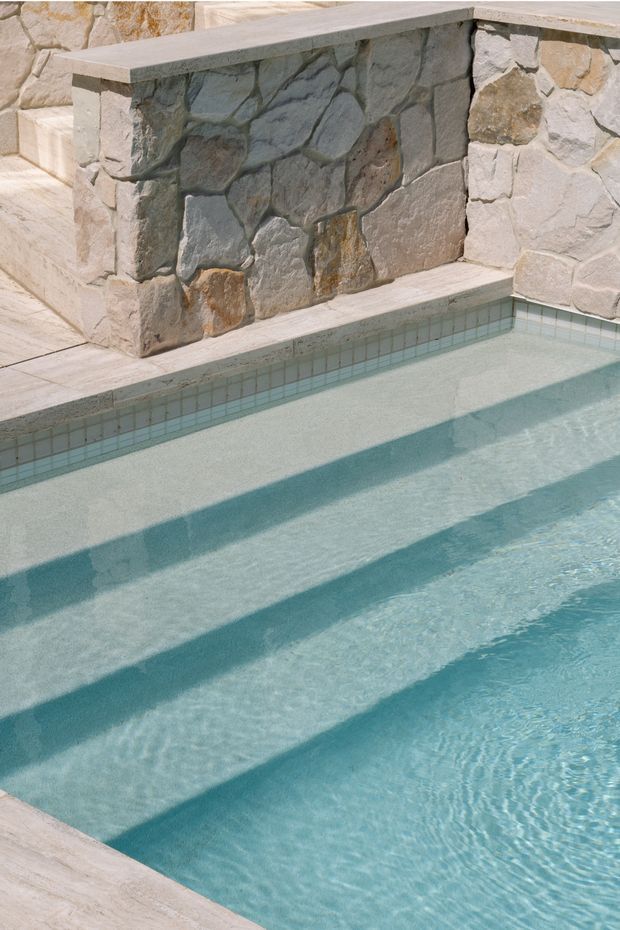 Close-up view of a pale turquoise swimming pool with broad submerged steps and strong diagonal shadows. The pool is edged with small light blue-green square tiles and pale stone paving, with a rustic irregular-stone retaining wall and raised stone ledge at the far side.