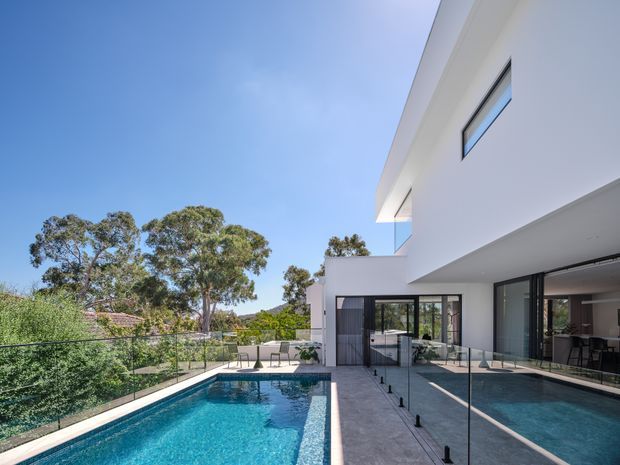 Exterior view of a contemporary white multi-level residence with strong cantilevered roof and balcony forms, extensive floor-to-ceiling glazing, and a narrow lap-style swimming pool beside a paved terrace. Glass balustrades and reflective glazing connect the indoor living spaces with the pool area, while mature trees are visible beyond the property.