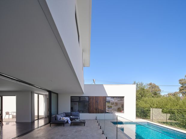 Contemporary multi-level residence viewed from a covered outdoor terrace, with crisp white rendered walls, broad roof overhangs, dark-framed floor-to-ceiling glazing, and a timber-clad section around the kitchen or living area. A glass balustrade borders the terrace and overlooks a rectangular swimming pool, while a minimalist outdoor seating area sits beside the open-plan interior.