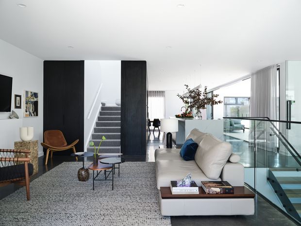 Bright contemporary open-plan living area with a large light sectional sofa, textured grey rug, low coffee tables, and a television wall. A black timber-lined stair enclosure creates a strong architectural feature, with stairs leading upward behind the living space. Full-height glazing, slim curtains, and a glass balustrade overlook a lower-level void or stairwell. The palette is predominantly white, black, grey, and light timber with selected warm brown furniture accents.