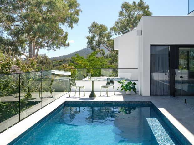 Bright contemporary rooftop terrace with a rectangular swimming pool, clear glass balustrade, white rendered building walls, and black-framed glazing. Outdoor dining furniture and potted planting sit beside the pool, with eucalyptus trees and a wooded hillside visible beyond the property.