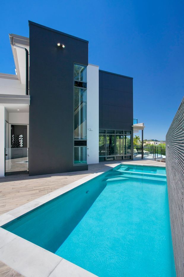 Exterior view of a contemporary multi-level residence centered on a long turquoise swimming pool. The building combines dark charcoal cladding or rendered wall planes with crisp white sections, extensive floor-to-ceiling glazing, and a narrow vertical glazed stairwell. A pale timber-look deck and light paving surround the pool, with a textured boundary wall along the right side. The image is photographed in bright daylight under a clear blue sky.