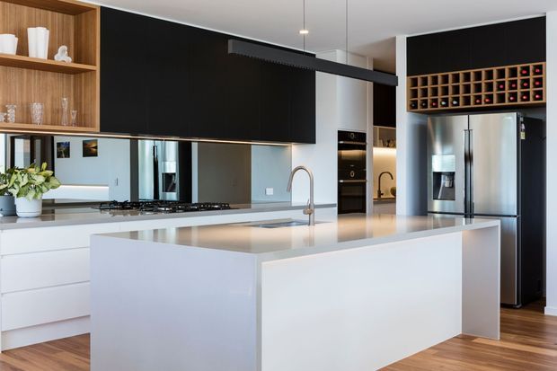 A contemporary open-plan kitchen dominated by a large white island with an integrated sink and curved tap. The space features white handleless cabinetry, a reflective dark splashback, timber open shelving, built-in black appliances, a stainless-steel refrigerator, timber flooring, and a wall-mounted wine rack. A long black linear pendant light runs above the island.