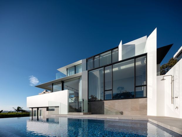 A striking multi-level contemporary residence with crisp white geometric forms, extensive floor-to-ceiling glazing, dark metal framing, and projecting roof planes. The house opens directly onto a reflective swimming pool, with a minimal outdoor shower and landscaped edge visible under a clear blue sky.