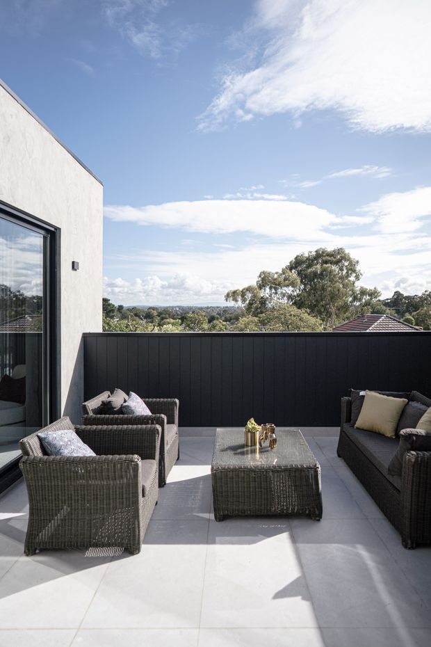 A bright rooftop or elevated outdoor terrace furnished as a lounge, with woven dark-grey armchairs and a matching sofa arranged around a rectangular coffee table. Large pale grey floor tiles, a tall black privacy screen, and a light rendered wall with dark-framed glazing define the space. Beyond the screen are treetops and suburban rooftops under a blue sky.