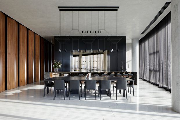 A dramatic contemporary dining area with a long dark dining table and multiple black upholstered chairs, positioned between tall timber wall panelling and a dark built-in kitchen or bar zone. Floor-to-ceiling sheer curtains line the glazed wall, while linear pendant lighting and polished pale flooring create a bright, refined interior.