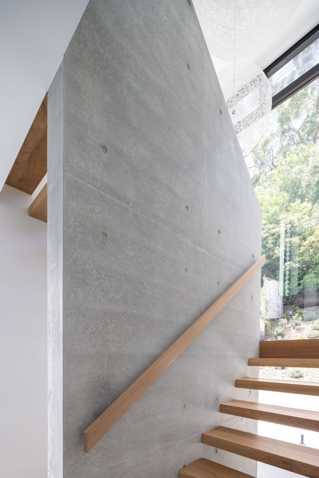 Interior architectural view of a modern open-riser staircase beside a tall exposed concrete wall. The staircase has warm timber treads and a matching timber handrail, with large windows bringing in natural light and views of trees. Exposed timber roof or ceiling elements are visible at the upper left, and a perforated spherical pendant light hangs near the glazing.
