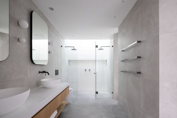 Bright contemporary bathroom with large light-grey tiled walls and floor, twin white vessel basins on a timber-fronted vanity, rounded black-framed mirrors, minimalist black tapware, open glass shower enclosure, and sleek wall-mounted towel rails.