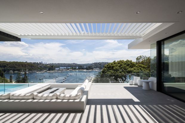 A contemporary waterfront outdoor terrace overlooking a marina, with a swimming pool, built-in white lounge seating, glass balustrades, and large glazed doors. A retractable or louvred roof structure casts strong linear shadows across the pale terrace flooring, creating a bright, open indoor-outdoor entertaining space.