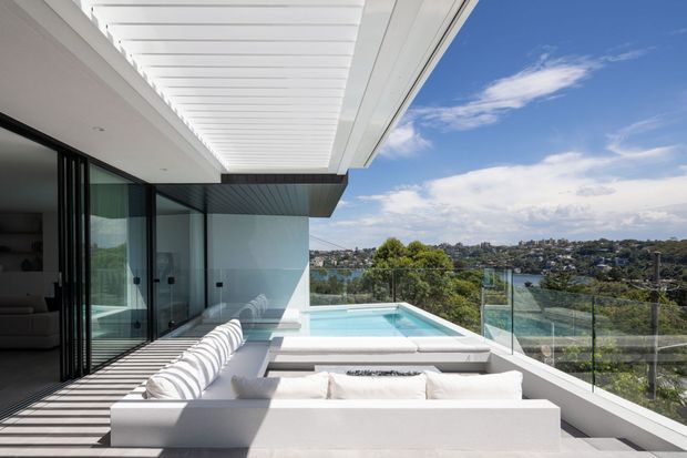 A bright contemporary rooftop terrace overlooking a leafy waterfront suburb. The space features a white built-in outdoor lounge, a glass balustrade, a small turquoise swimming pool, large sliding glass doors, and a white slatted overhead canopy creating strong linear shadows.