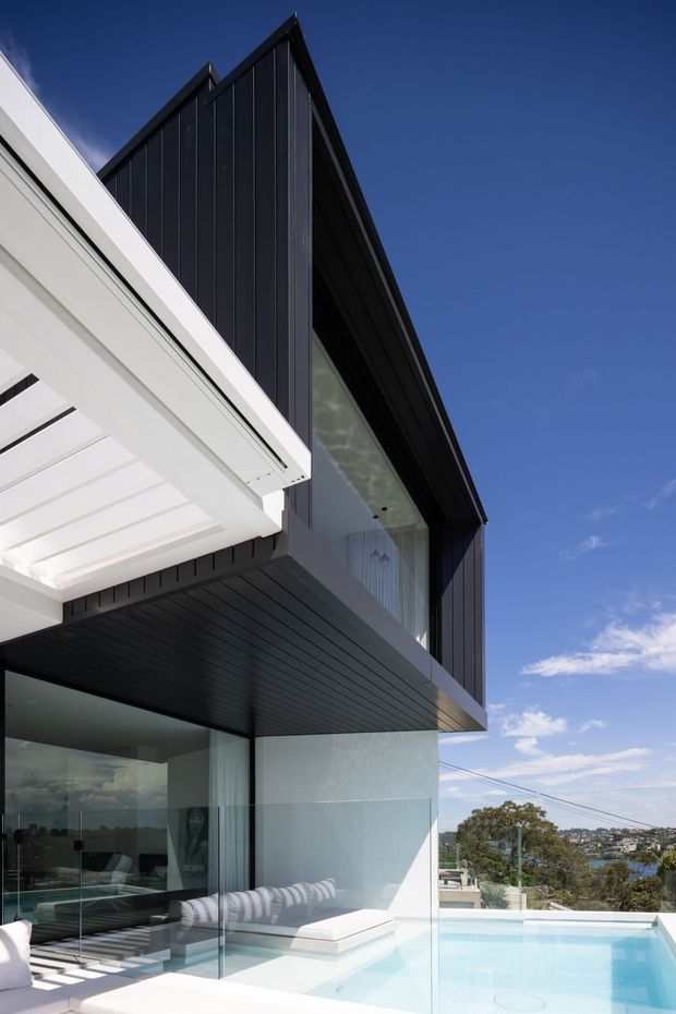 A contemporary multi-level residential exterior with a dramatic steep gable roof, dark vertical cladding, white structural elements, extensive glazing, and a cantilevered upper floor. A bright pool terrace with frameless glass balustrades sits below, overlooking a suburban landscape under a clear blue sky.