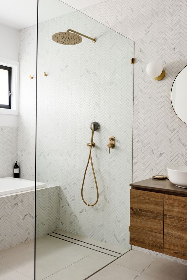 A refined contemporary bathroom featuring a frameless glass walk-in shower, white herringbone wall tiles, large pale floor tiles, brushed brass shower fittings, and a floating dark timber vanity with a white vessel basin. A freestanding or built-in bathtub is partially visible beside a black-framed window, with a round wall mirror and globe wall light adding decorative detail.