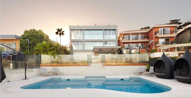 A landscaped residential pool area viewed from the pool deck, with a large swimming pool in the foreground, glass balustrades and lounge seating, dark pod-like outdoor chairs, and several multi-storey homes in the background. The scene has a warm, resort-like contemporary feel.