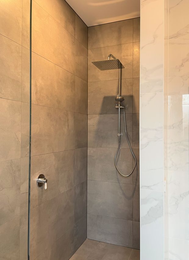 A modern walk-in shower enclosure with large warm grey-beige wall and floor tiles, a frameless glass screen, and brushed metal rain shower and handheld fixture. A white marble-look tiled wall is visible at the right edge. The space is warmly lit and minimalist.