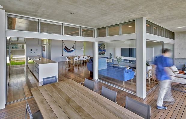 Open-plan contemporary home interior viewed from a dining area, with a large timber dining table in the foreground, a white kitchen island to the left, and a living room visible through extensive internal glazing. The space features exposed concrete ceilings, warm timber flooring and decking, white structural frames, clerestory-style internal windows, and abundant natural light. A person is present but is not a defining feature.