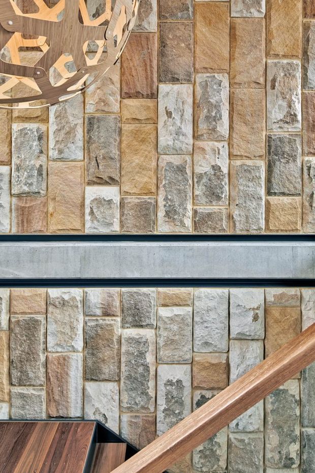 Close-up architectural interior detail showing a stairway with a timber handrail and dark timber stair treads, set against a prominent textured wall clad in irregular rectangular natural stone tiles. A decorative timber pendant or sculptural light is partially visible at the upper left, and a horizontal grey metal or concrete structural band crosses the wall.