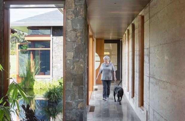 A long, sheltered residential hallway or covered passage with polished grey stone flooring, warm timber-lined ceiling, timber-framed glazing and doors, and exposed rough stone and concrete-block walls. The passage connects to a landscaped courtyard visible through the glazing, with a person and dog walking through the space. The architecture has a grounded, natural-material character with strong indoor-outdoor connection.