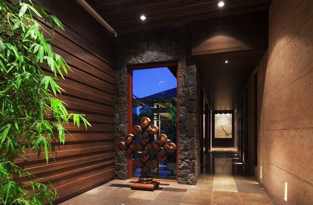 A dramatic, dimly lit interior corridor or entry hall with dark horizontal timber wall cladding and a matching slatted ceiling. A rough dark stone surround frames a glazed opening, beside a sculptural wooden artwork. The corridor extends toward a framed artwork at the far end, with warm recessed spotlights, polished stone or tile flooring, and a textured warm-toned wall. Bamboo-like planting adds an organic accent near the entrance.